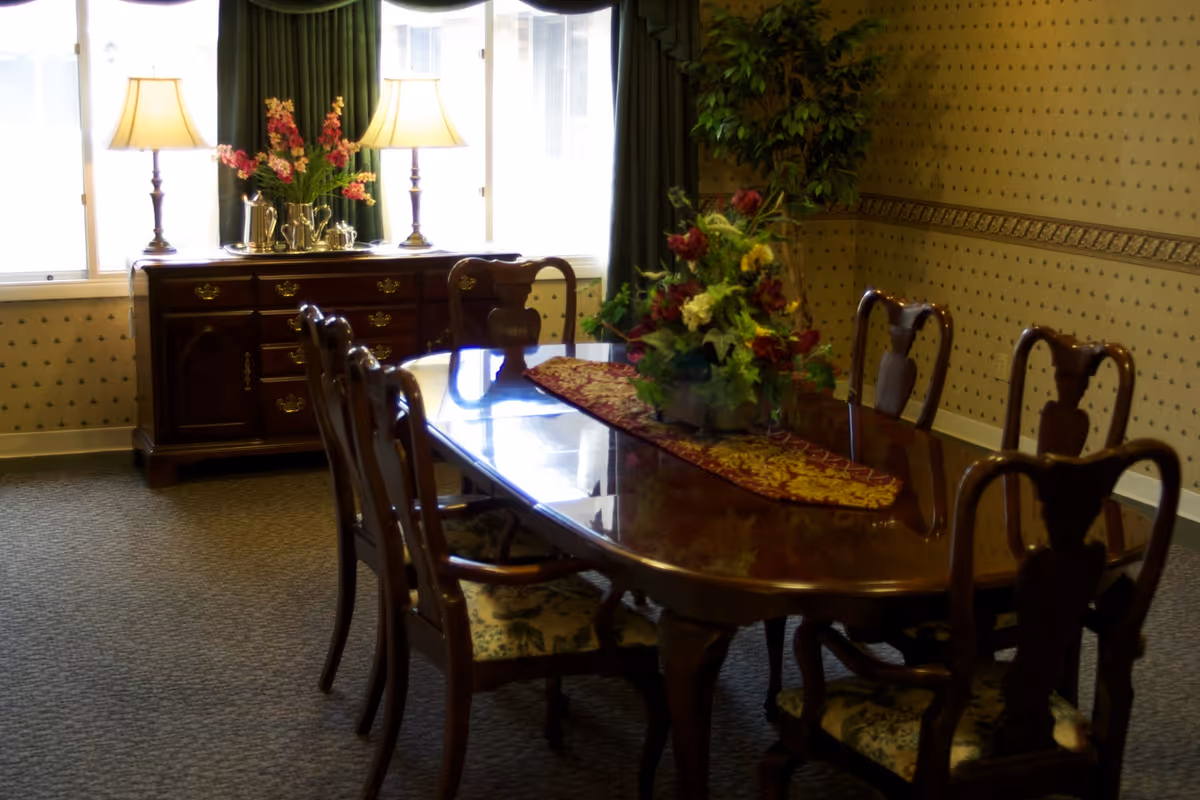 A traditional dining room with a polished wooden dining table surrounded by six wooden chairs with floral upholstered seats. The table has a decorative runner and a floral centerpiece. In the background, there is a wooden sideboard with two lit table lamps and a silver tea set, positioned in front of a window with dark green curtains. The walls have patterned wallpaper and there is a large potted plant in the corner.