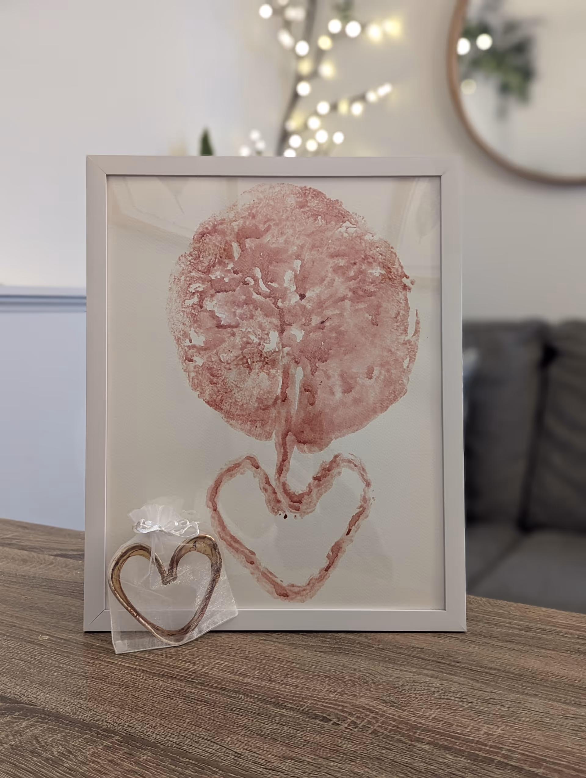 Framed pink watercolor artwork on a wooden table with a small heart pendant.