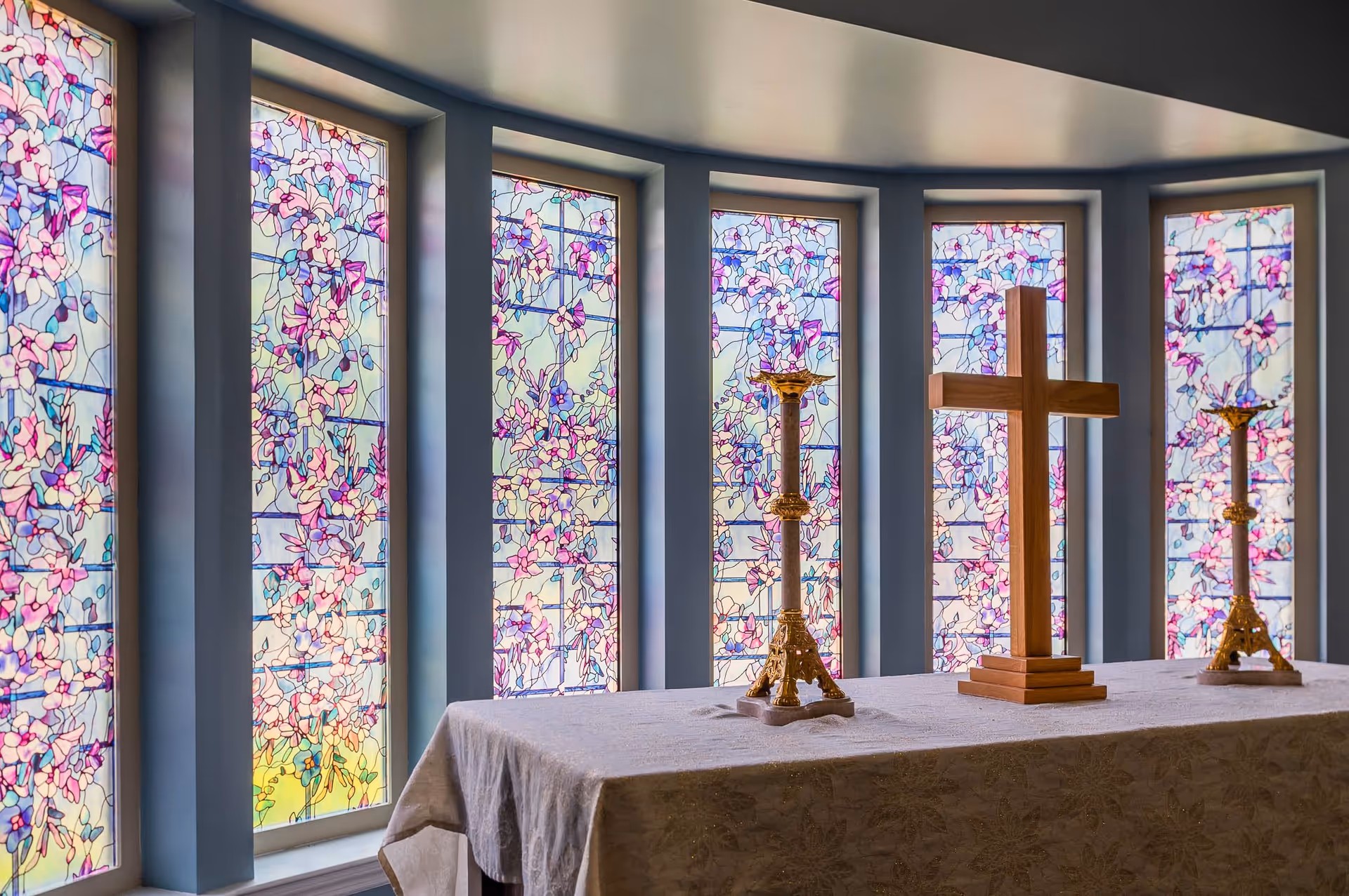 Interior view of a chapel with a table covered by a white cloth, holding a wooden cross and two ornate golden candlesticks. Behind the table are tall stained glass windows with floral patterns in shades of pink, purple, and blue.