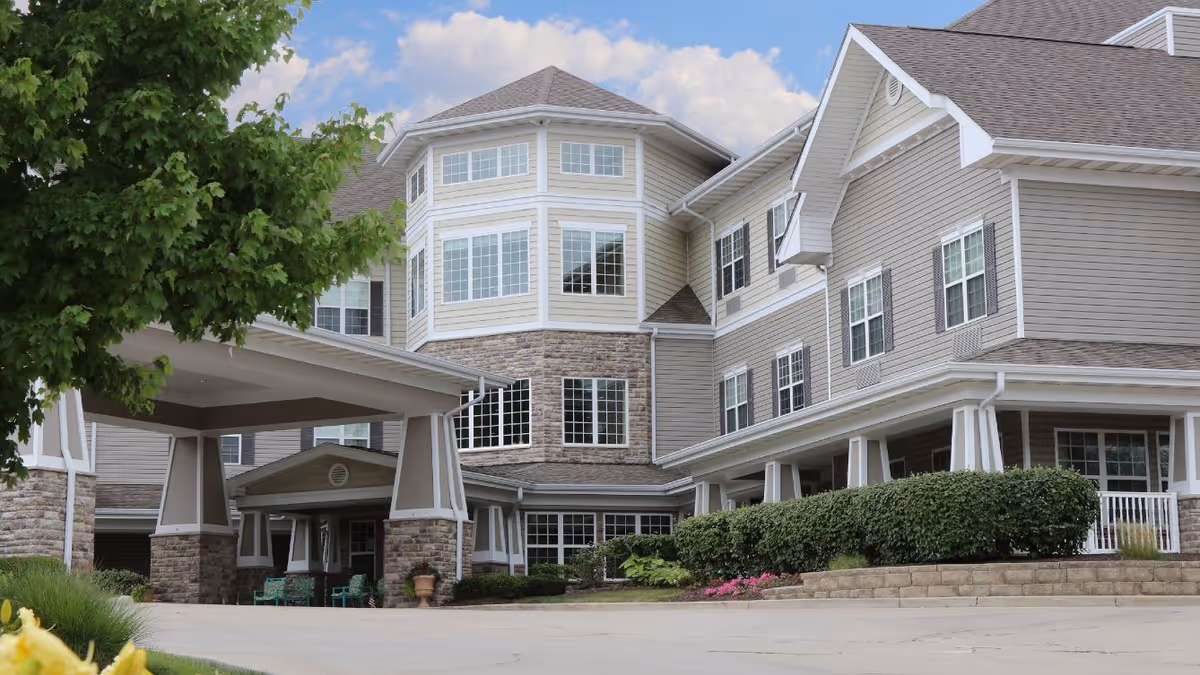 Exterior view of a multi-story senior living facility with beige siding and stone accents. The building features many windows, a covered entrance with pillars, manicured bushes, and a tree on the left side under a partly cloudy sky.