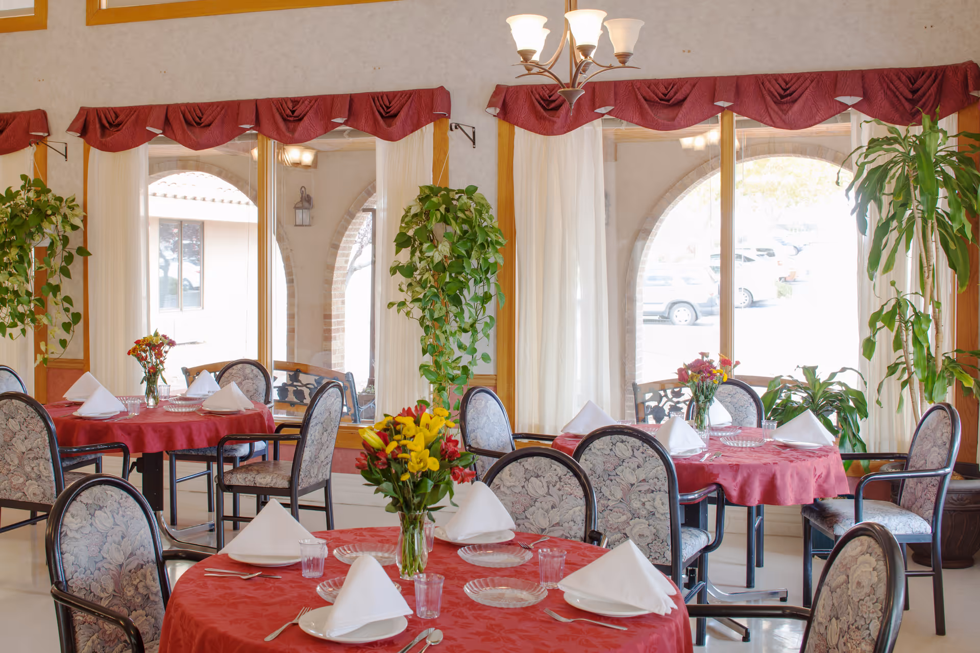 Bright dining room with round tables set with red tablecloths, white napkins, floral centerpieces and upholstered chairs in front of large arched windows.