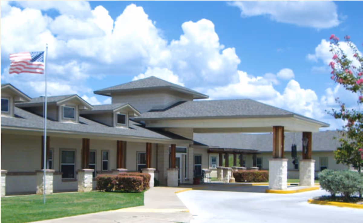 Front entrance of an assisted living building with a covered porte-cochère, flagpole, and a partly cloudy sky.