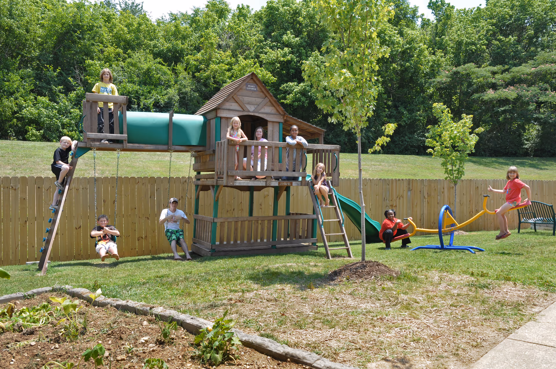 Children playing on a wooden playground set with a slide, swings, and a climbing ladder in a grassy fenced yard with trees and a garden bed nearby.