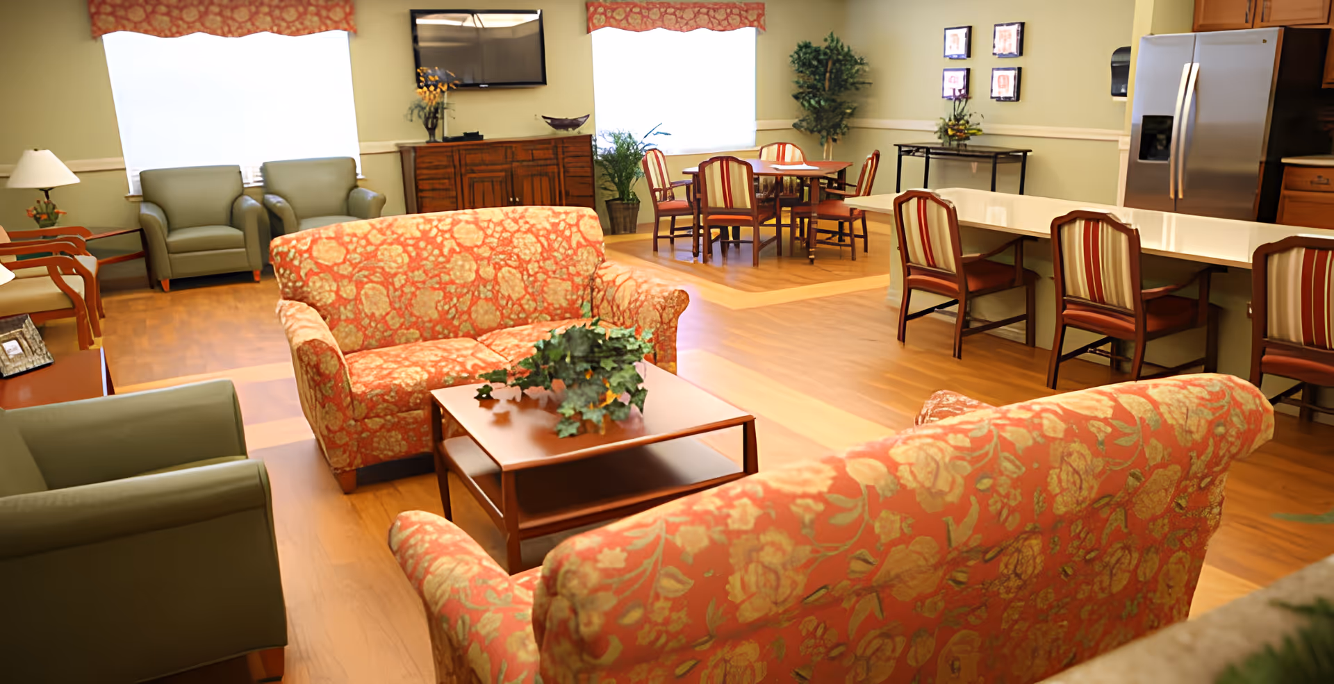 Open communal living area with floral sofas, armchairs, a coffee table, dining table and kitchenette in the background.