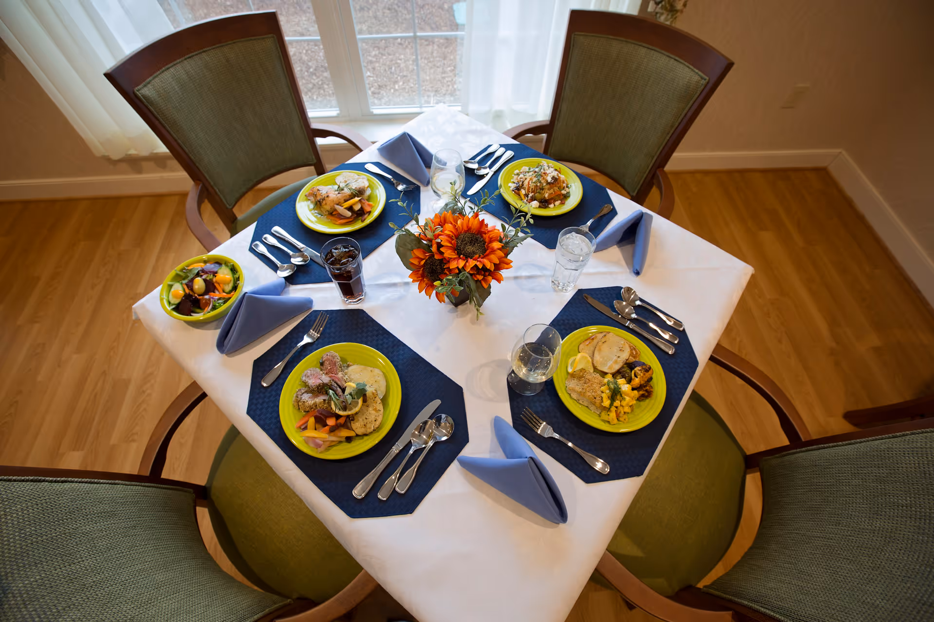 A dining table set for four with green cushioned chairs around it. The table is covered with a white tablecloth and blue placemats, each setting has a plate with a meal, silverware, a glass of water or soda, and a folded blue napkin. A small vase with orange flowers is in the center of the table. The setting is near a window with sheer white curtains.