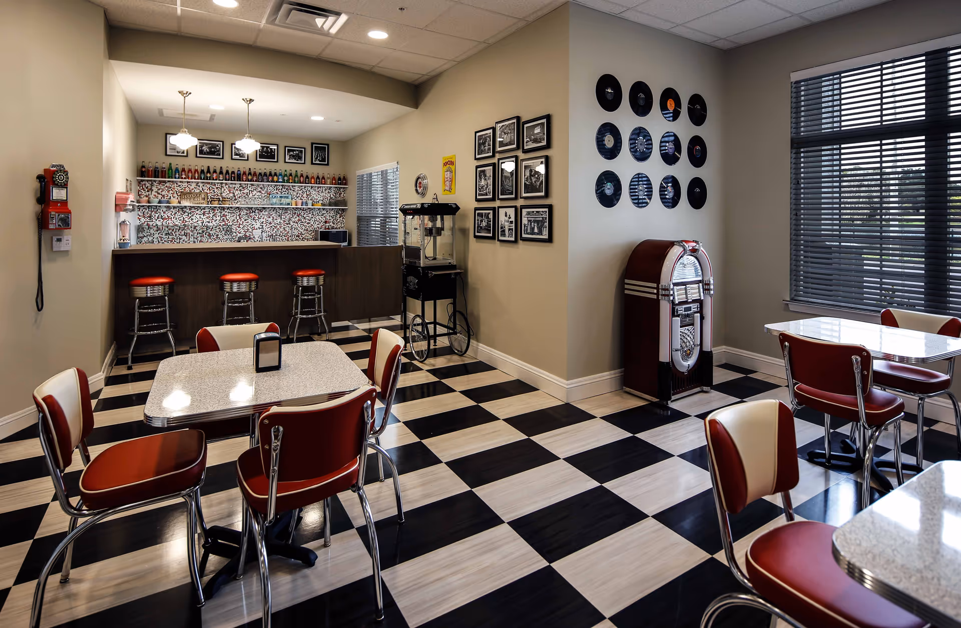 Retro-style dining area with black-and-white checkered floor, red diner chairs and tables, a counter with stools, a jukebox and wall records.