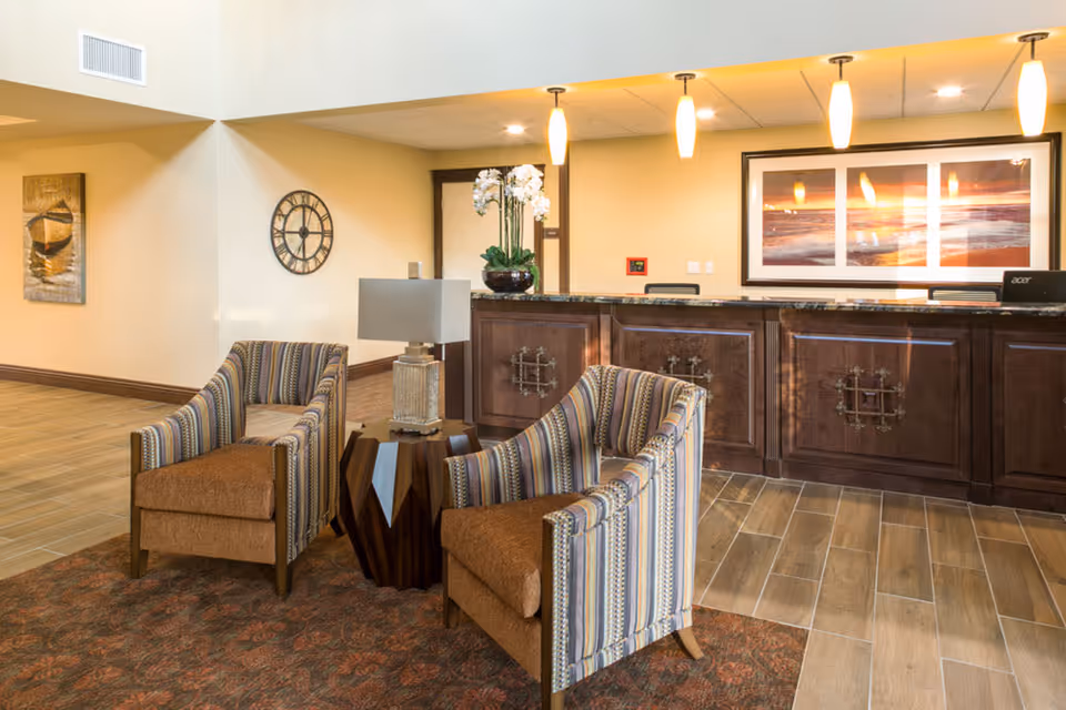 A cozy seating area with two striped armchairs and a small wooden table with a lamp, situated in front of a dark wood reception desk with granite countertop. The reception area is well-lit with hanging pendant lights and features a large framed picture on the wall behind the desk. The floor is tiled with a wood-like pattern and a patterned rug is under the chairs.