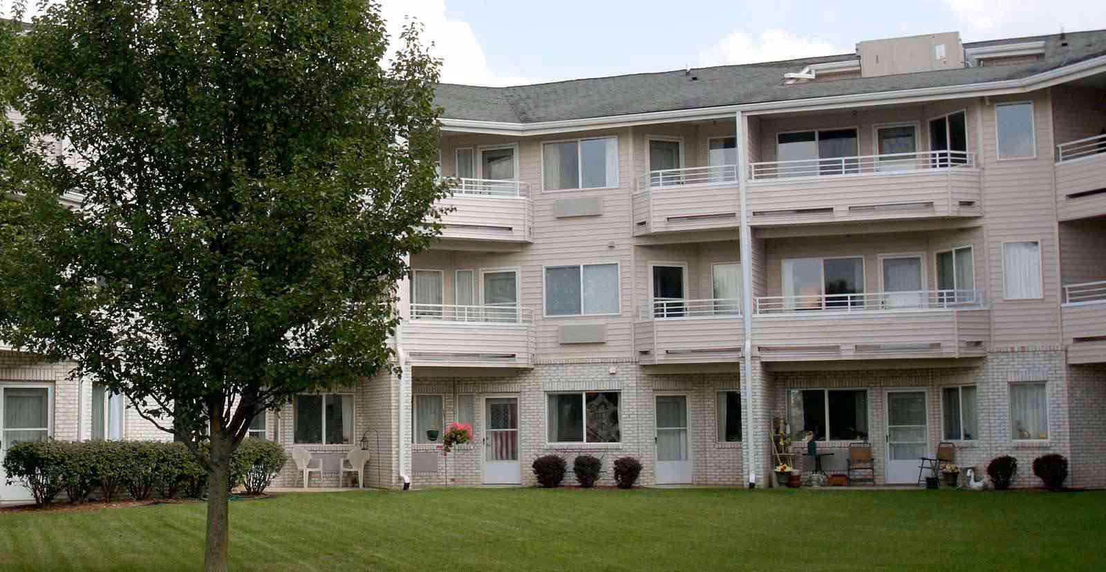 Three-story senior living building with balconies, a grassy lawn and a tree in front.