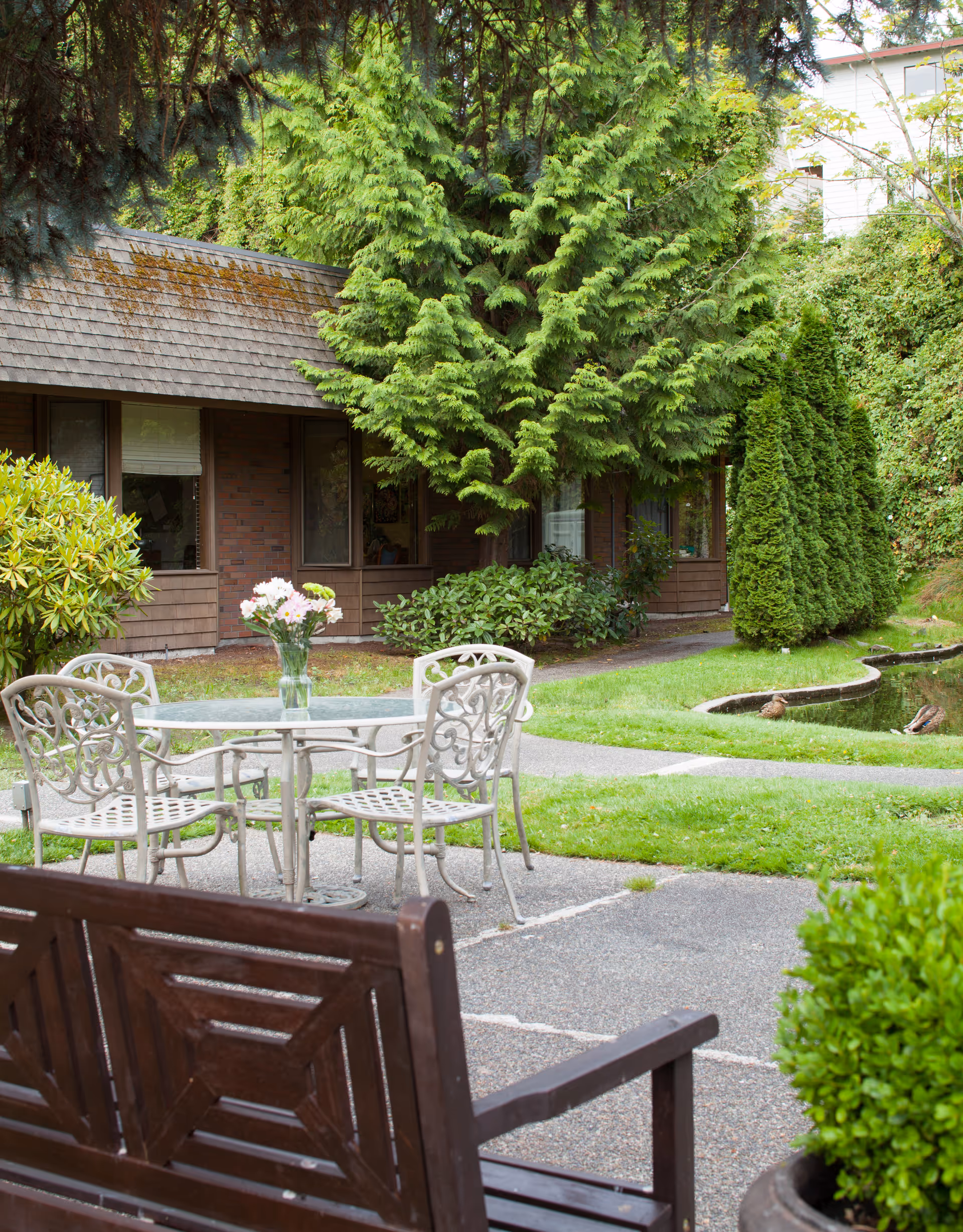Outdoor garden area at Ballard Center featuring a glass-top metal table with four chairs, a wooden bench in the foreground, lush green trees and bushes, a small pond with ducks, and a building with large windows in the background.