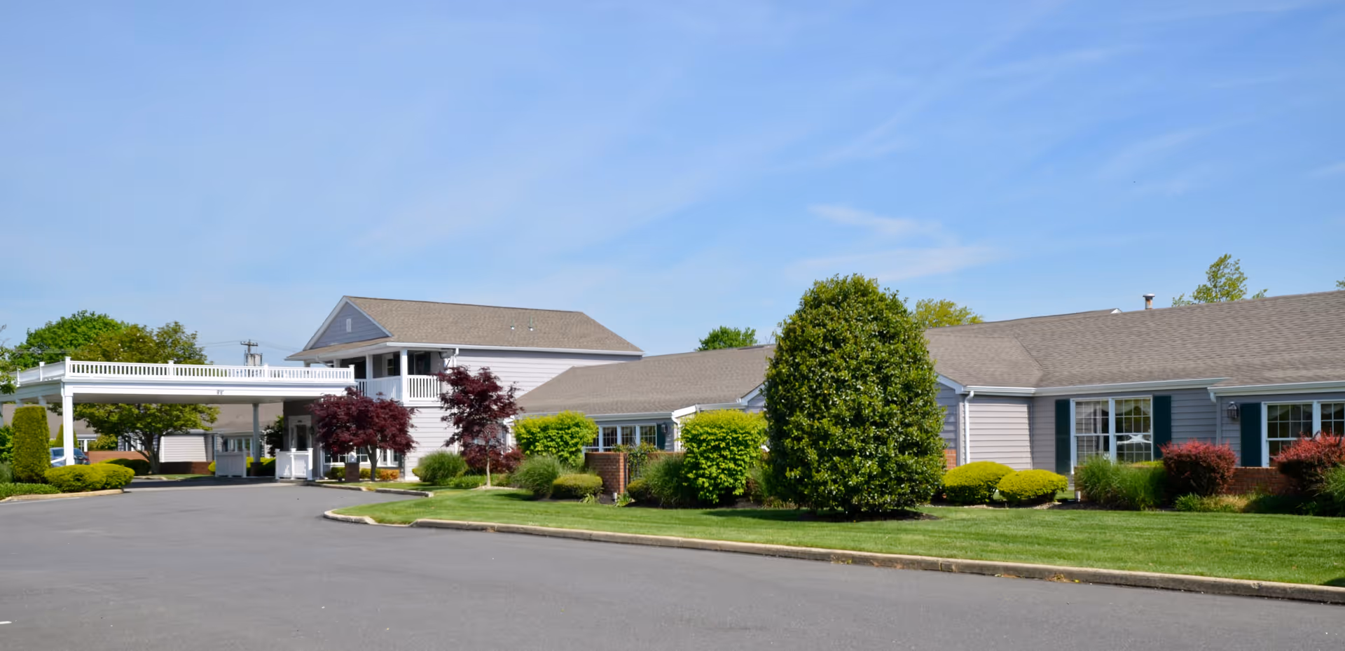 Front exterior of a single-story senior living building with a covered entryway, shrubs, and a paved driveway under a blue sky.