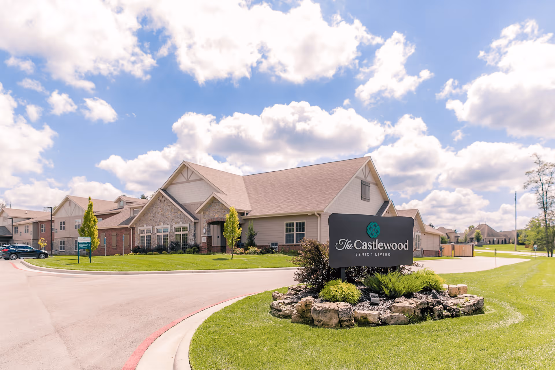 Exterior view of The Castlewood Senior Living facility on a sunny day with a partly cloudy sky. The building has a combination of stone and siding with a pitched roof. In the foreground, there is a landscaped area with a sign that reads 'The Castlewood Senior Living'.