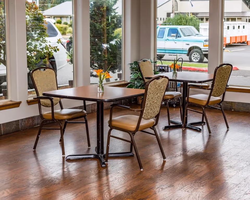 Dining area with tables and chairs by large windows inside a senior living facility.