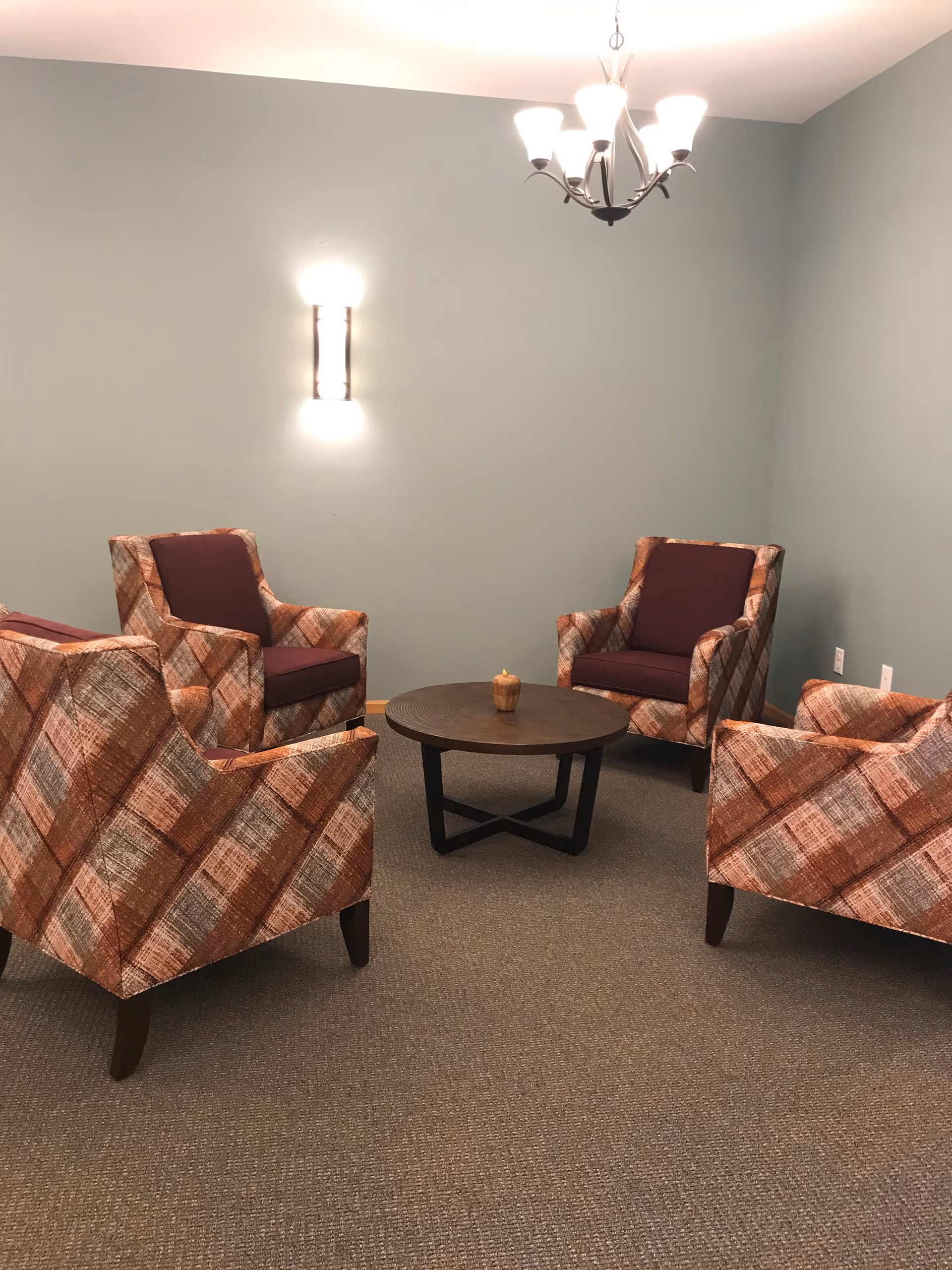 A small seating area with four patterned armchairs arranged around a round wooden coffee table with a small decorative item on it. The room has light gray walls, a wall-mounted light fixture, and a ceiling chandelier with five lights. The floor is carpeted in a neutral tone.