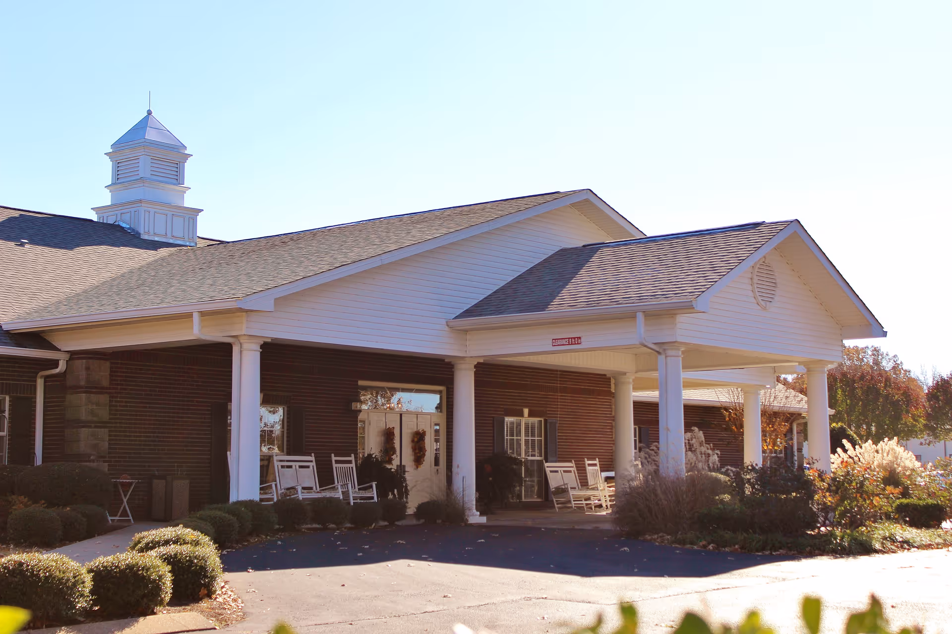 Front exterior view of Sugar Creek Senior Living facility showing a covered entrance with white columns, rocking chairs on the porch, and well-maintained landscaping with bushes and ornamental grasses.