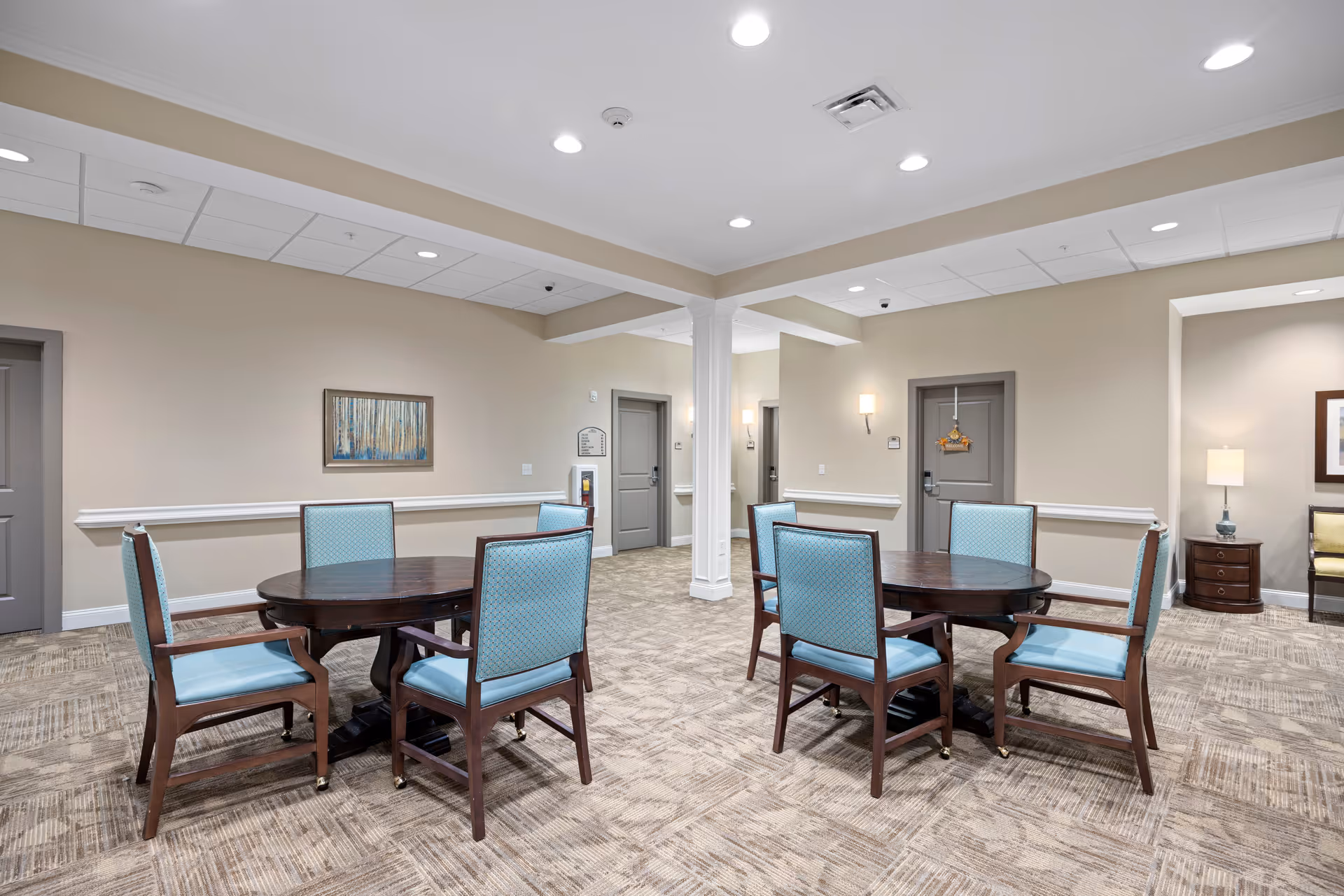 A bright communal room with two round wooden tables surrounded by light-blue upholstered chairs and neutral decor.
