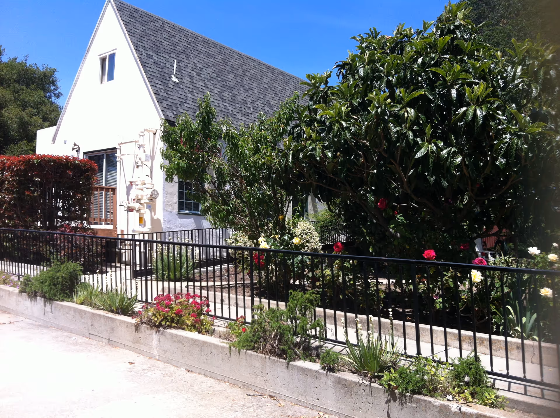 Front exterior of a white gable-roof house with a fenced garden and flowering bushes under a clear blue sky.
