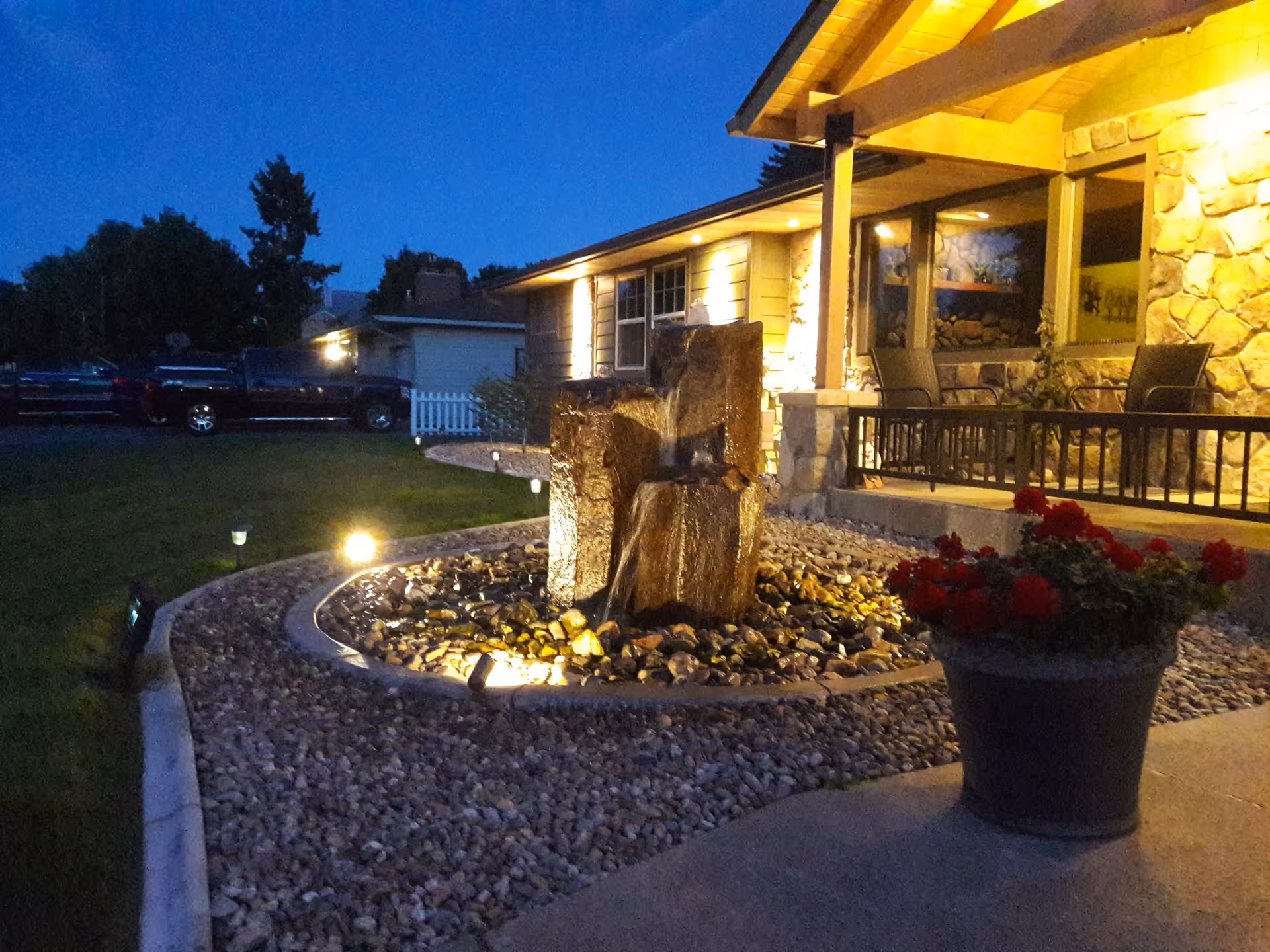 Evening view of the exterior of a retirement home building with warm lighting. In the foreground, there is a stone water fountain surrounded by rocks and a large flower pot with red flowers. The building has a porch with chairs and stone walls, and there are trees and parked vehicles in the background.