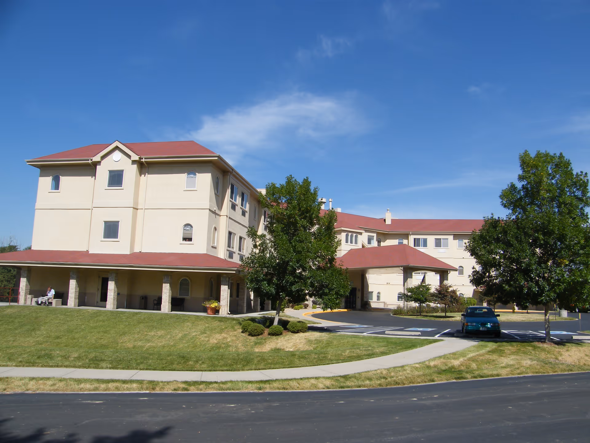 Exterior view of a three-story assisted living facility building with beige walls and a red roof under a clear blue sky. There is a covered entrance with a driveway, a few trees, a parked car, and a well-maintained lawn with a sidewalk in front.