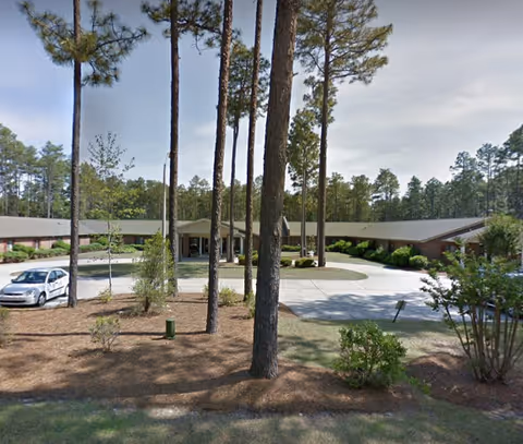 Magnolia Gardens senior living facility exterior with circular driveway, tall trees, and a low single-story building.
