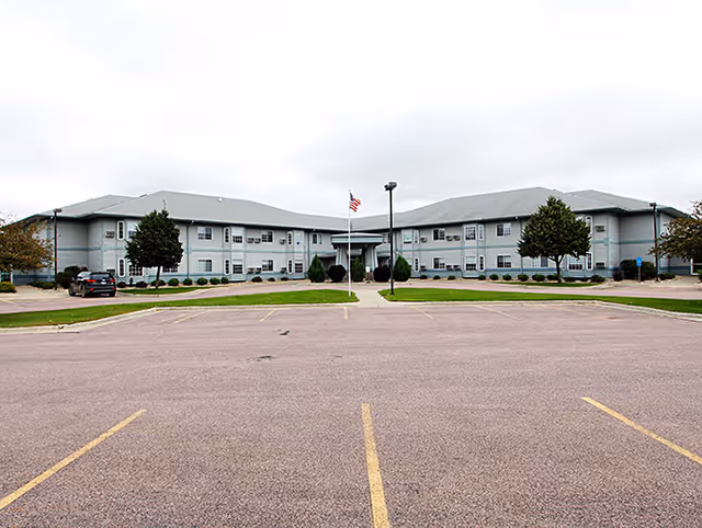 Front exterior view of a two-story senior living facility building with a gray roof and light-colored walls, an American flag on a flagpole in front, a few trees, and an empty parking lot in the foreground under a cloudy sky.