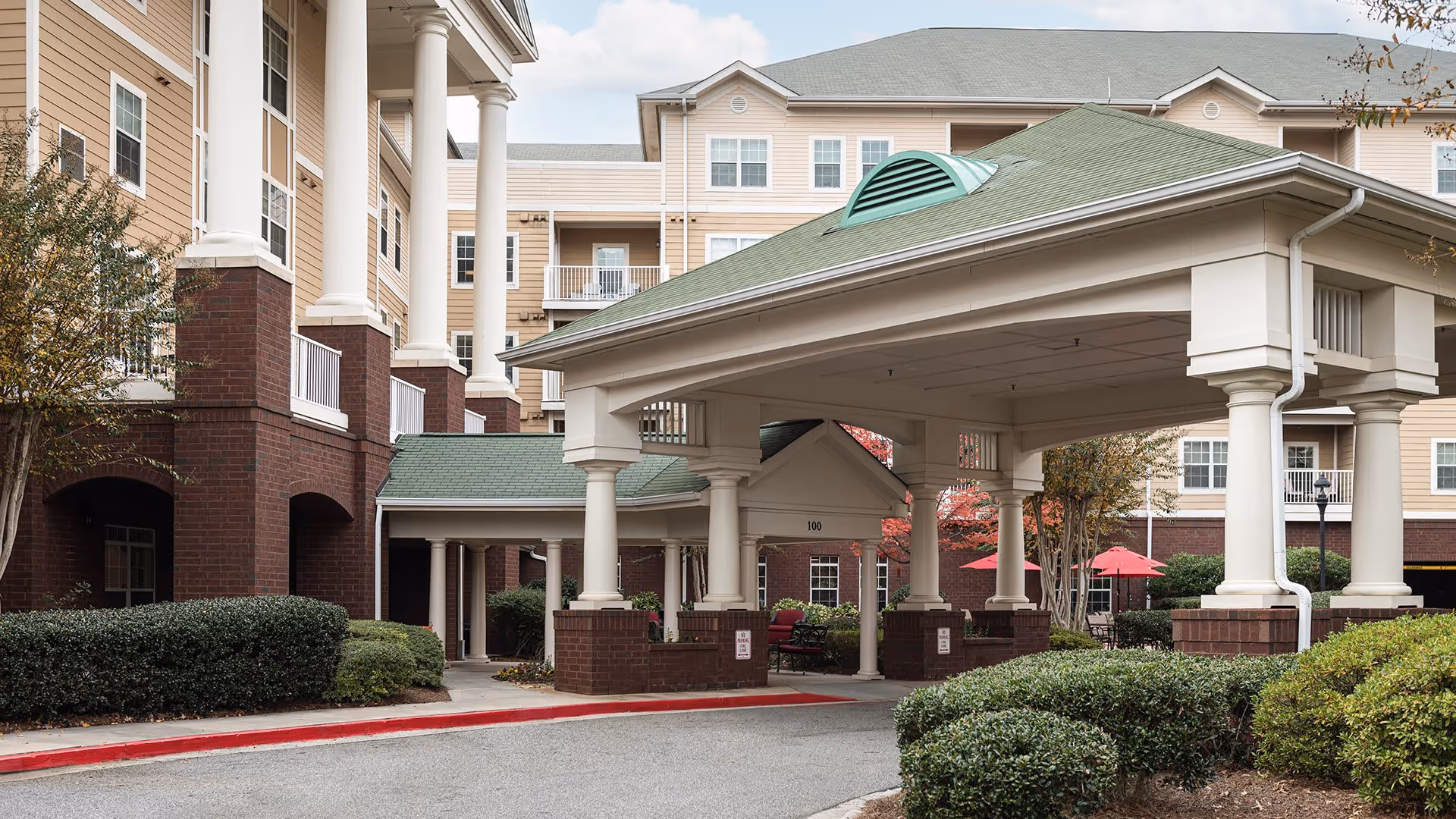 Entrance area of a senior living facility with a covered drop-off zone supported by white columns and brick bases. The building has beige siding with white trim and multiple windows. There are bushes and small trees around the driveway, and red umbrellas are visible in the background near a seating area.