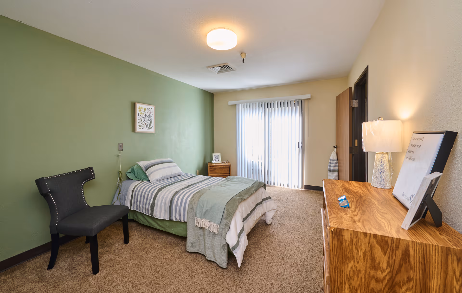 A bedroom with a single bed against a green accent wall, striped bedding, a black chair, a wooden dresser with a lamp and framed pictures, beige carpet, and vertical blinds covering a sliding glass door.
