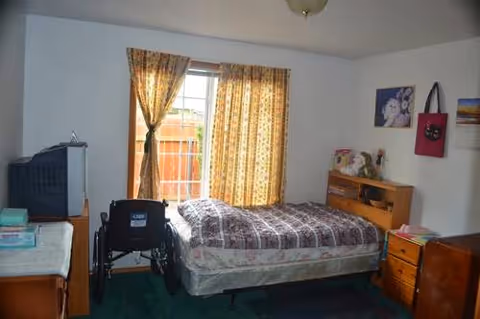 A bedroom with a bed, plaid quilt, curtains, dresser, and a wheelchair by the window.