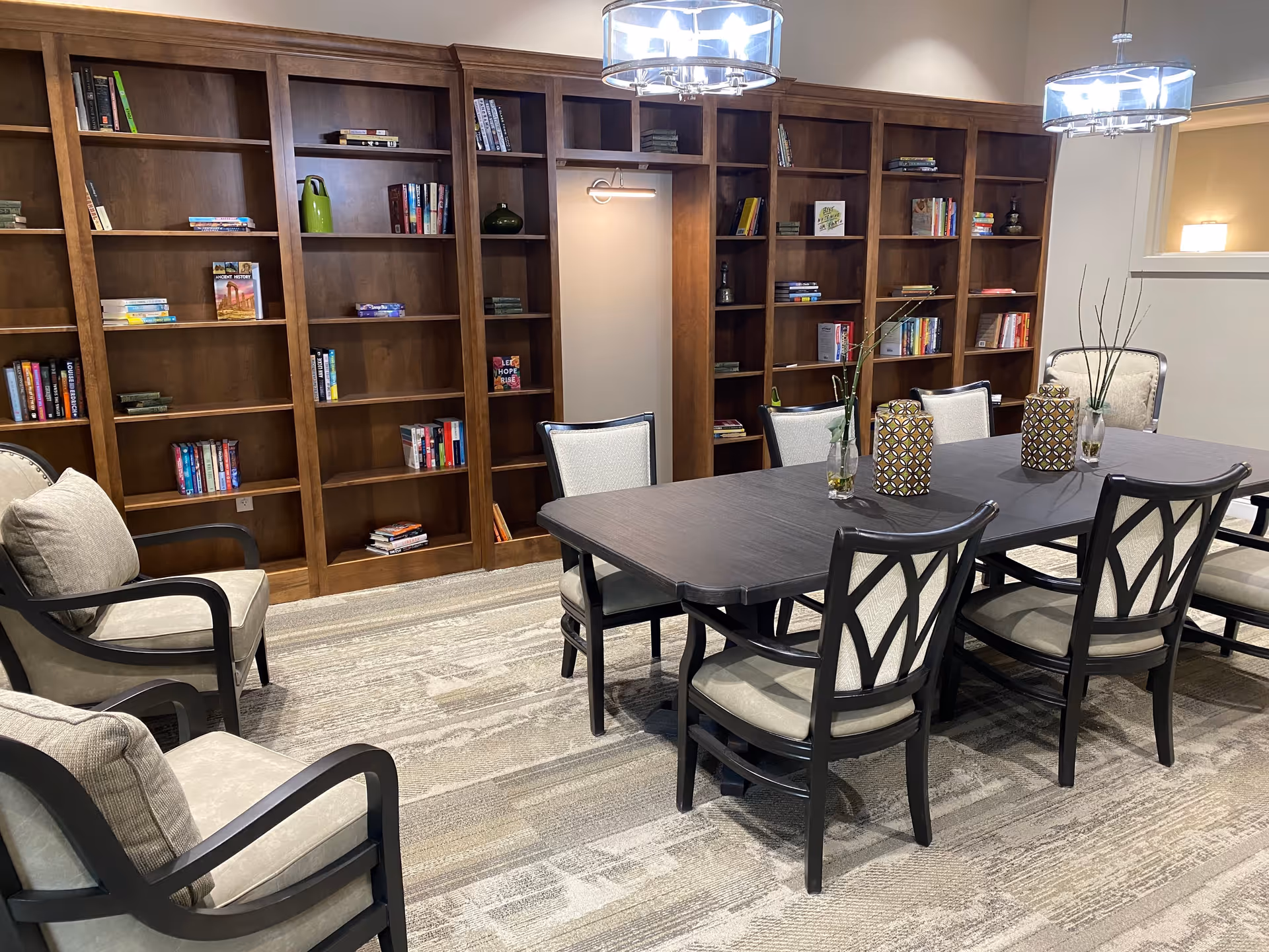 A well-lit communal dining and reading room with a long dark table surrounded by chairs and built-in wooden bookshelves along the back wall.