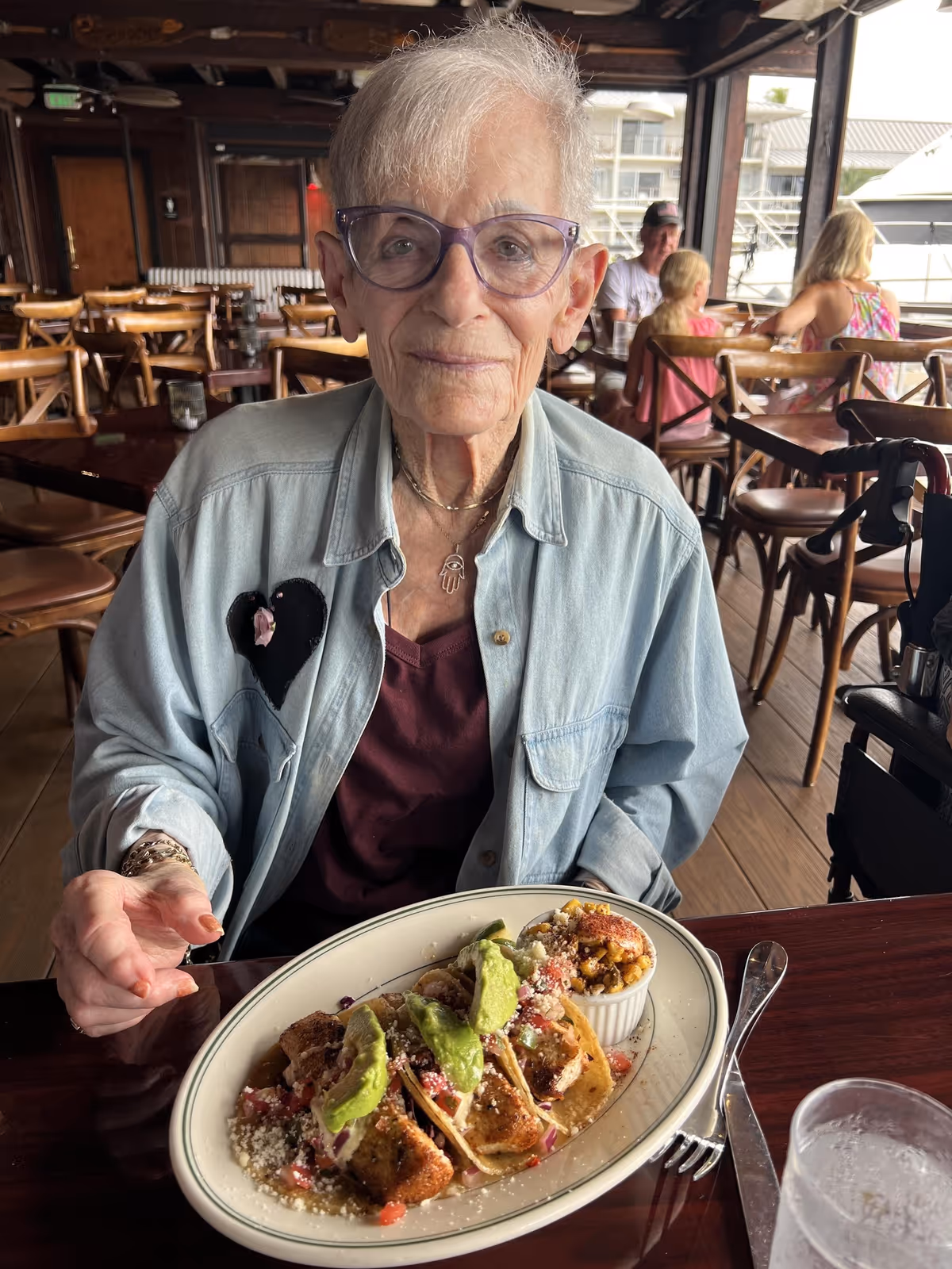 An elderly woman with short white hair and purple glasses sits at a wooden table in a restaurant, smiling gently. She is wearing a light denim jacket with a black heart patch and a maroon top underneath. In front of her is a plate with three tacos topped with avocado slices and a small ramekin of corn. The restaurant has wooden chairs and tables, and other diners are visible in the background.