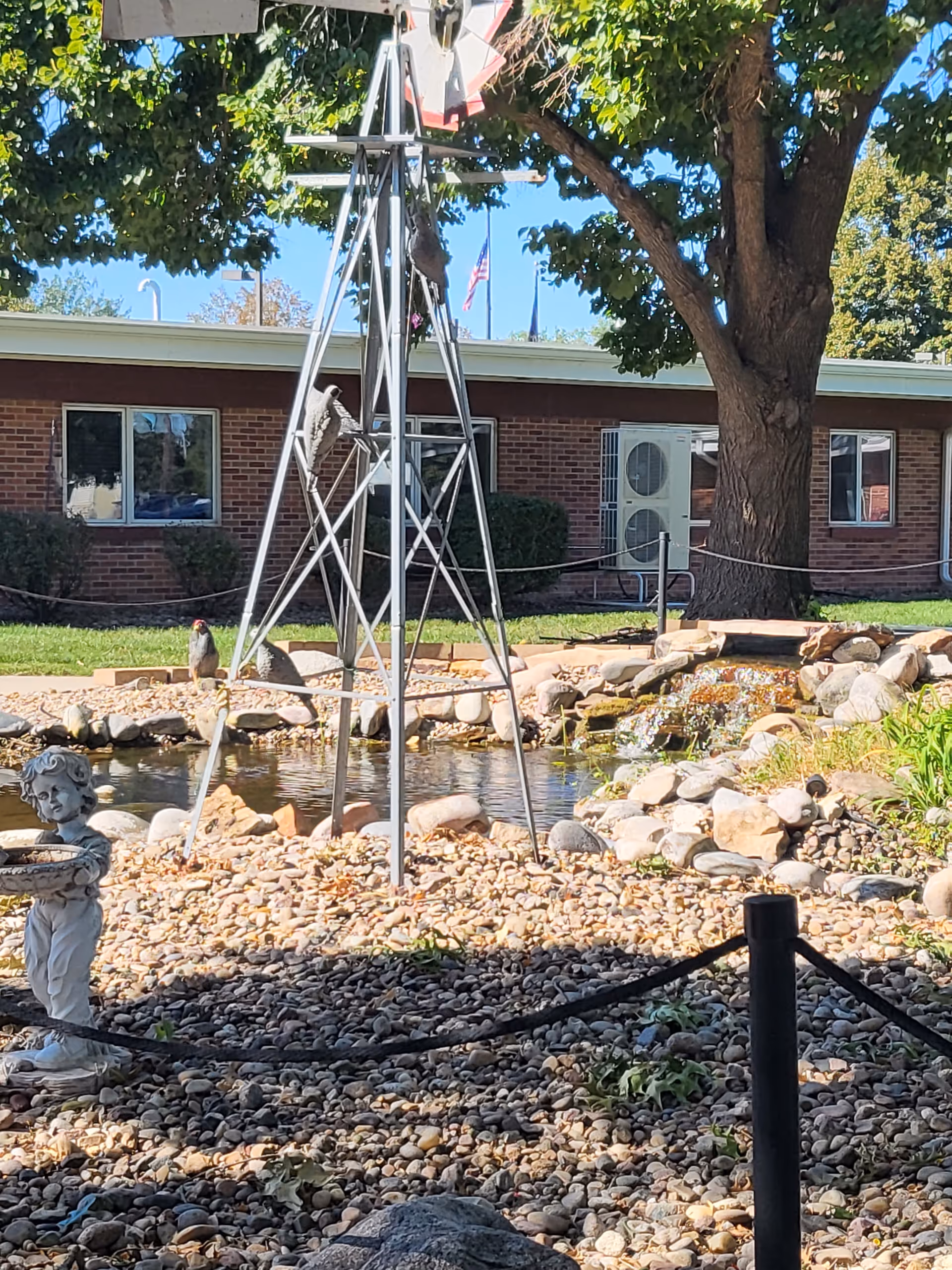 Outdoor rock garden with pond, statue, windmill sculpture, and brick building backdrop.