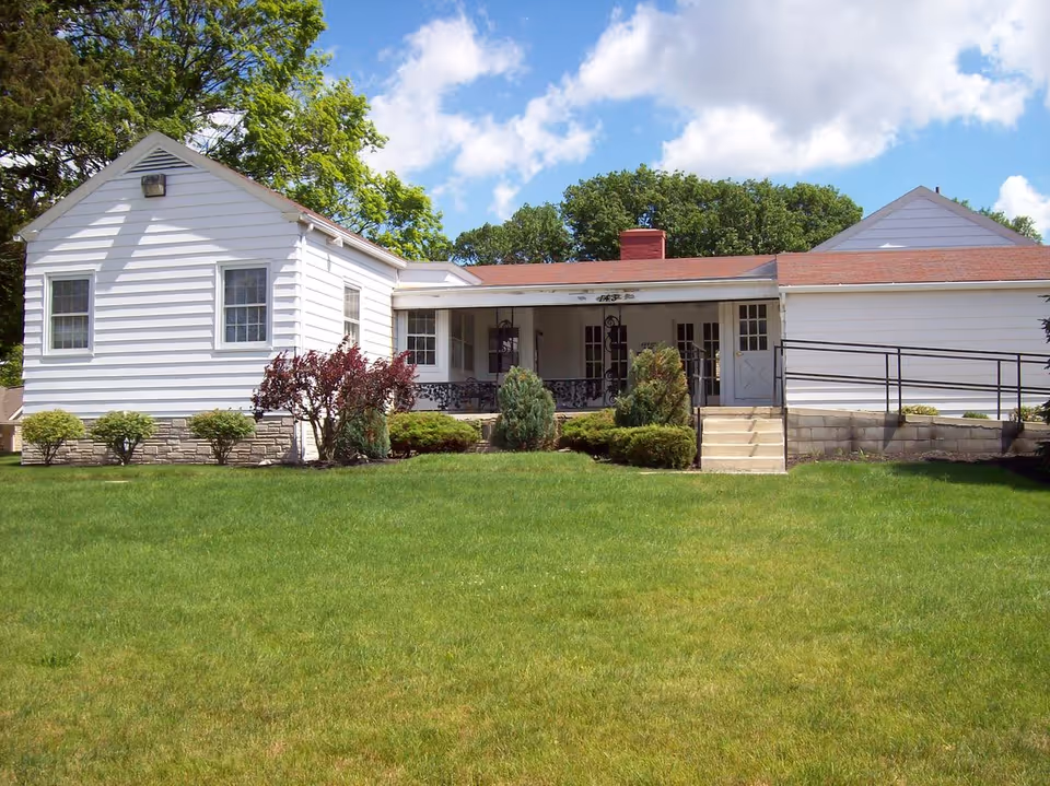 White single-story building with a front porch, ramp and steps set behind a wide green lawn under a blue sky.