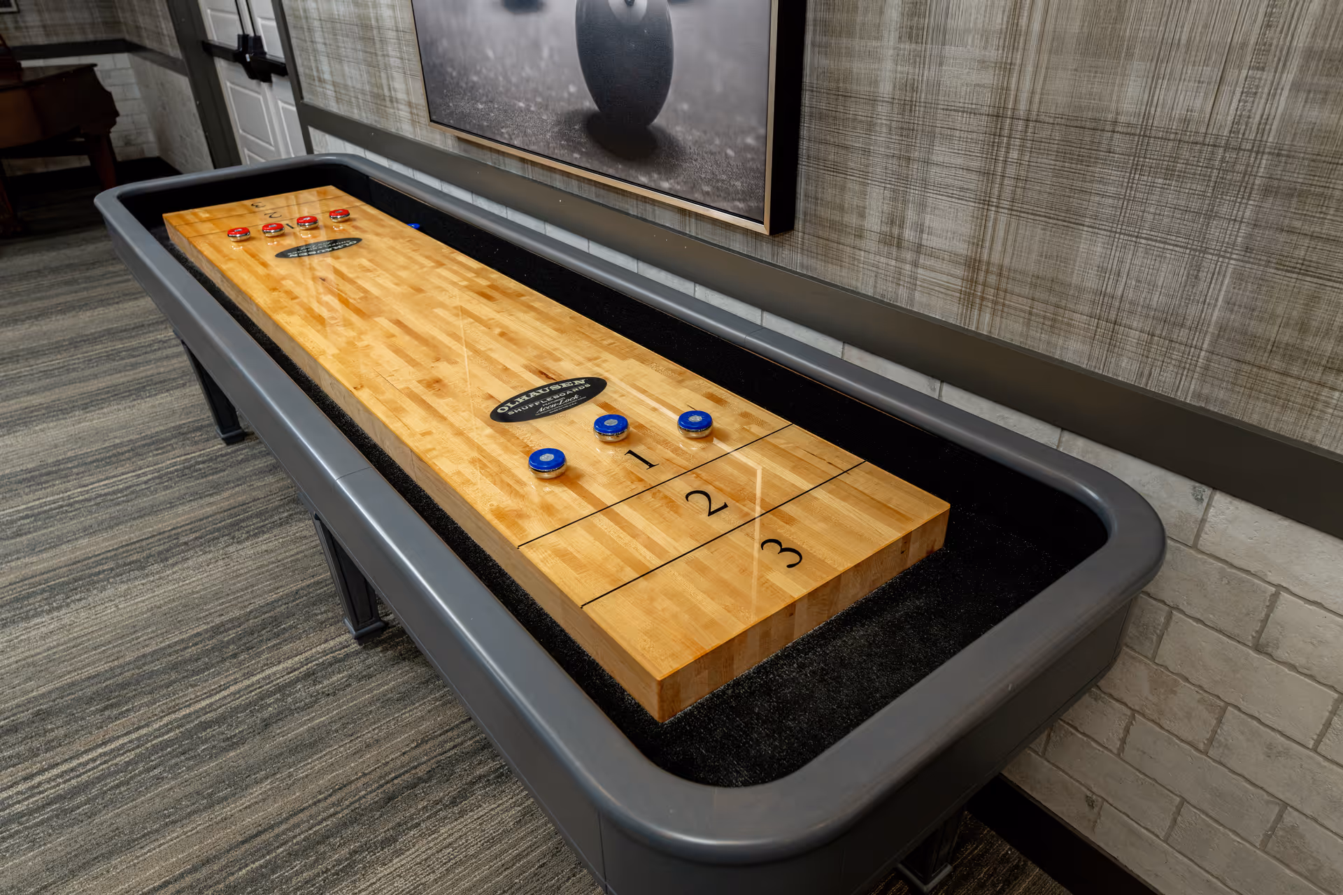 A wooden shuffleboard table sits in a patterned lounge area of a senior living facility.