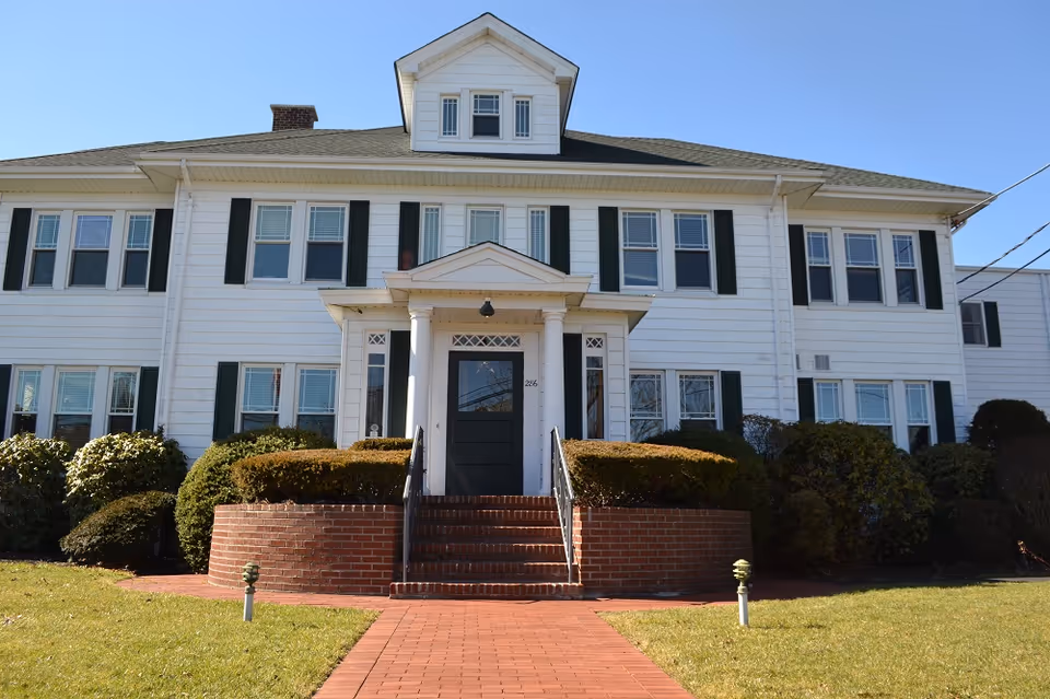 White two-story building with brick steps, columns, green shutters, and manicured shrubbery.