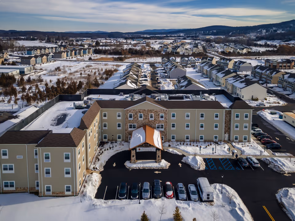 Aerial view of The Chelsea at Sparta senior living facility in a snowy landscape. The building is a three-story structure with a covered entrance and a parking lot in front. Surrounding the facility are residential houses and open fields with snow-covered ground and distant hills under a partly cloudy sky.