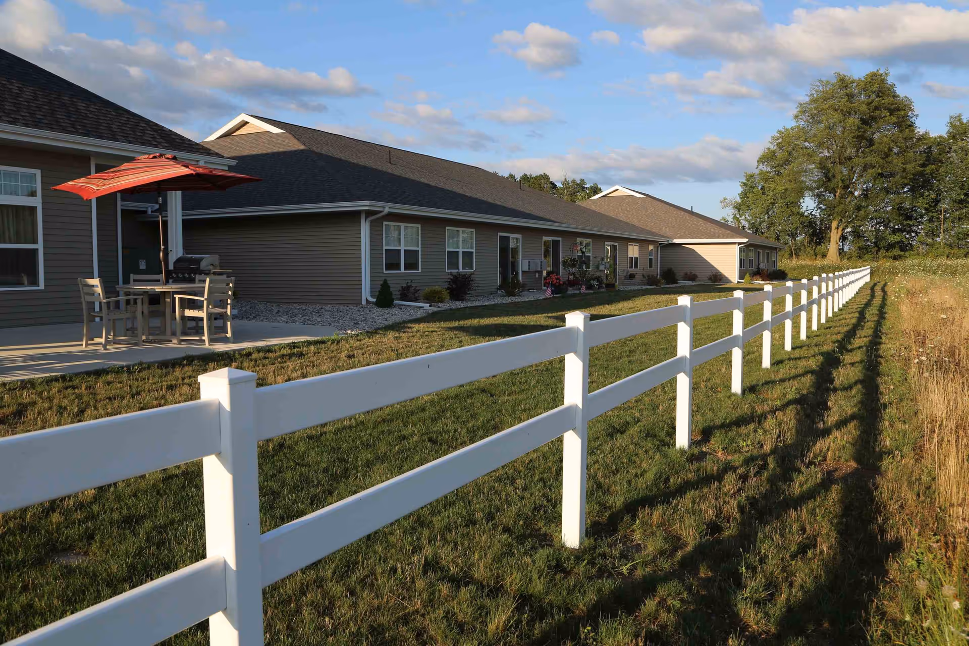 Single-story senior living building with a white fence, lawn, and a patio table with a red umbrella.