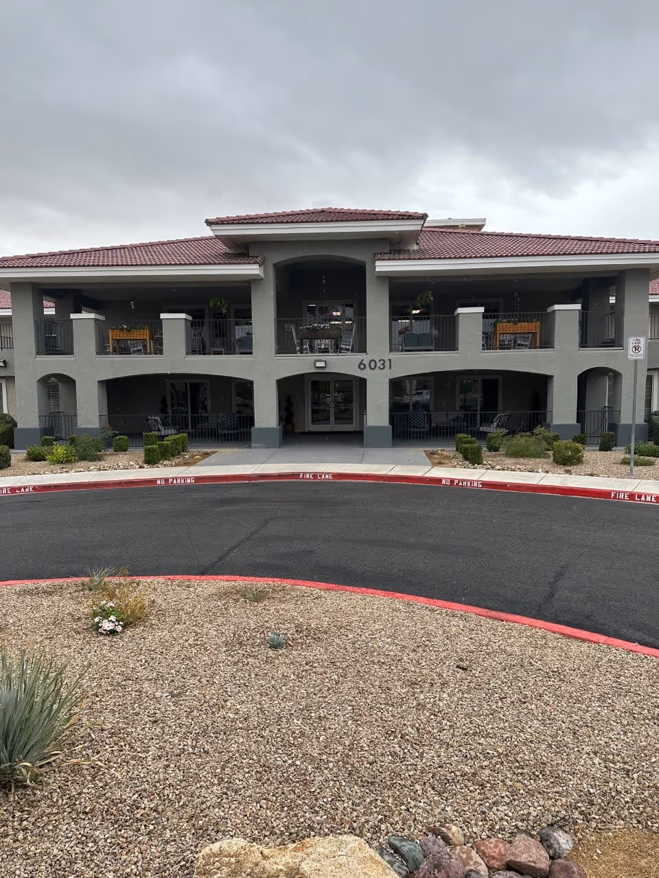 Front exterior view of a two-story building with a red-tiled roof and gray walls, featuring balconies on the upper floor and a covered entrance in the center. The building number 6031 is displayed above the entrance. The foreground shows a curved driveway bordered by red-painted curbs marked with 'FIRE LANE NO PARKING' and a landscaped area with gravel and sparse desert plants.