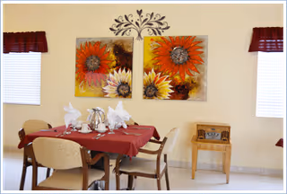 A dining area with a table covered by a red tablecloth set with silverware, napkins folded in a decorative manner, and a silver tea set. Four chairs surround the table. On the wall behind the table are two large sunflower paintings. There are two windows with red valances on either side of the paintings. A small wooden table with a vintage radio sits against the wall to the right.