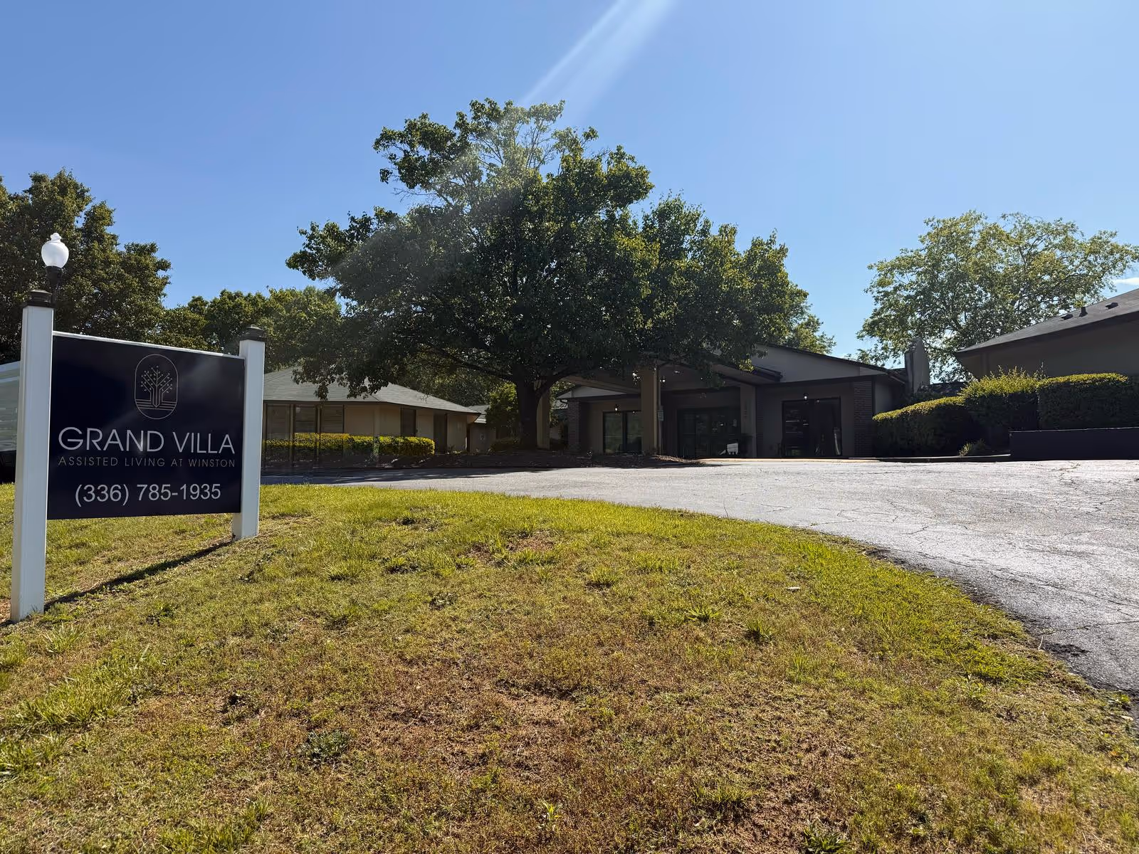 Front exterior of Grand Villa Assisted Living at Winston with a driveway, large tree, and a lawn sign displaying the facility name and phone number.