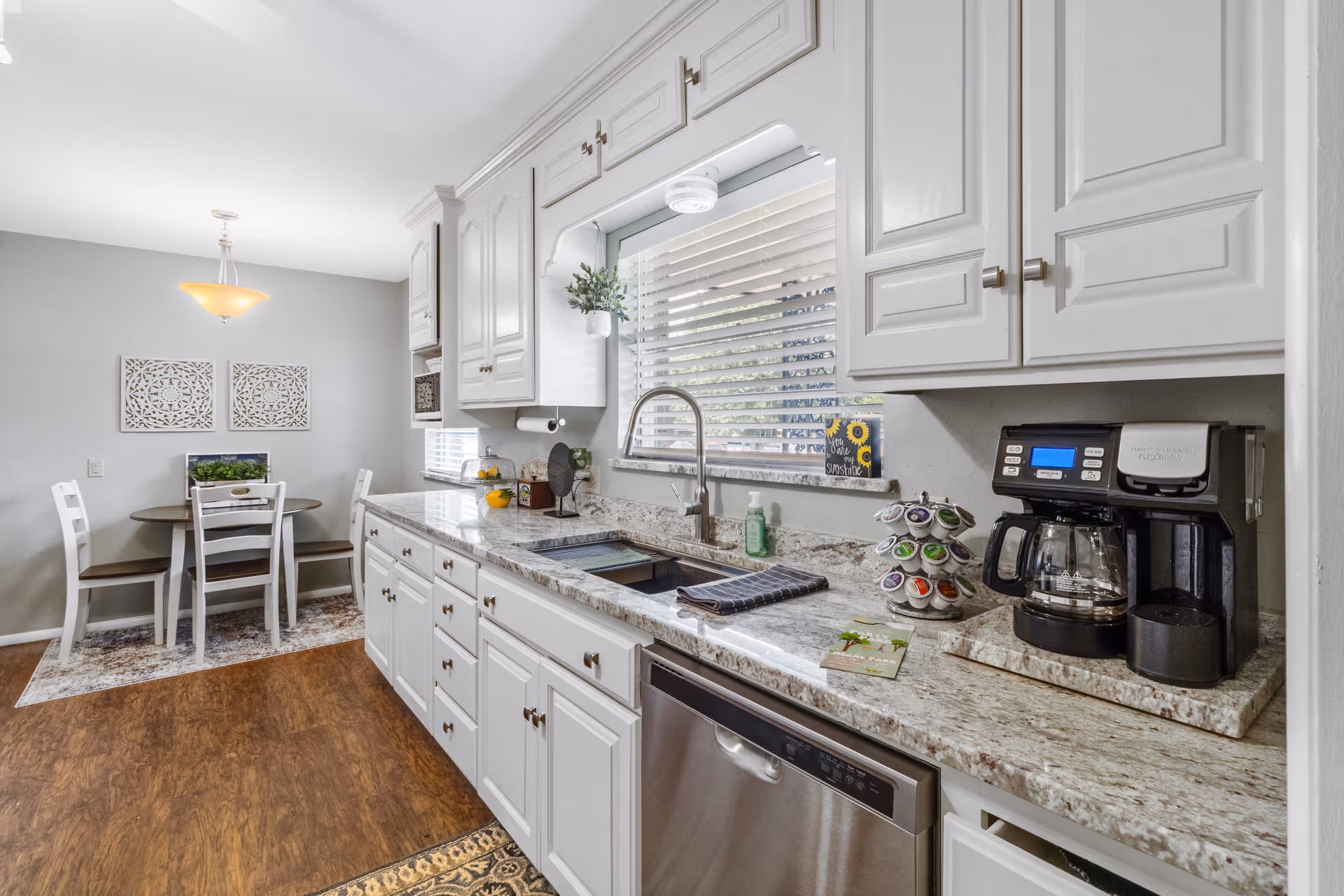 Bright white kitchen with granite countertops, stainless appliances, and a small dining nook in the background.