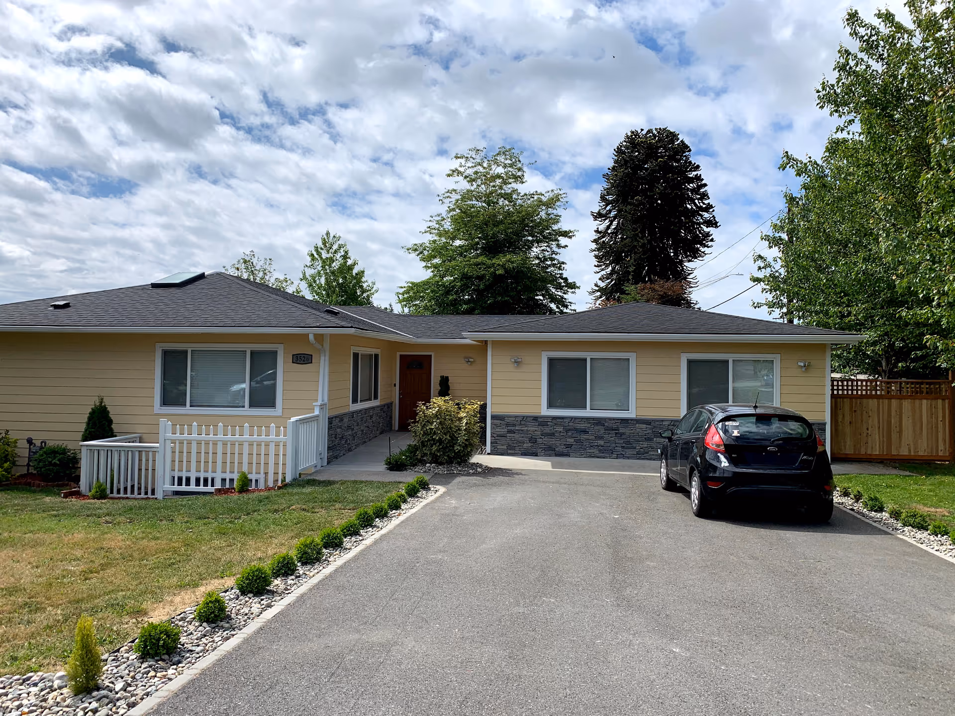 Single-story yellow senior living facility building with a gray roof, stone accents on the lower walls, and a driveway with a black car parked. The building is surrounded by green grass, small shrubs, and trees under a partly cloudy sky.