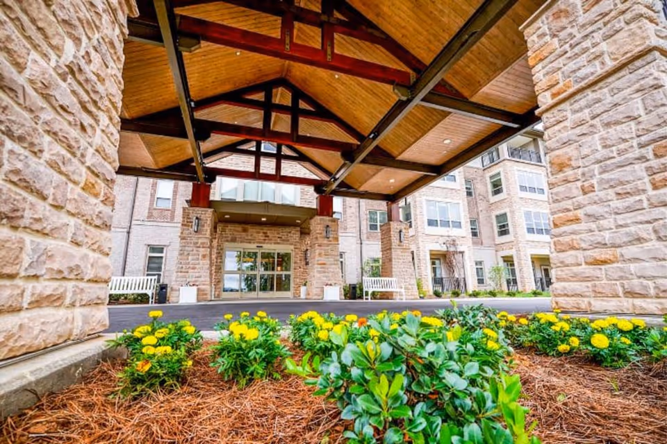 Covered porte-cochere entrance with timber beams and stone columns at the front of the building, with yellow flowers in the foreground.