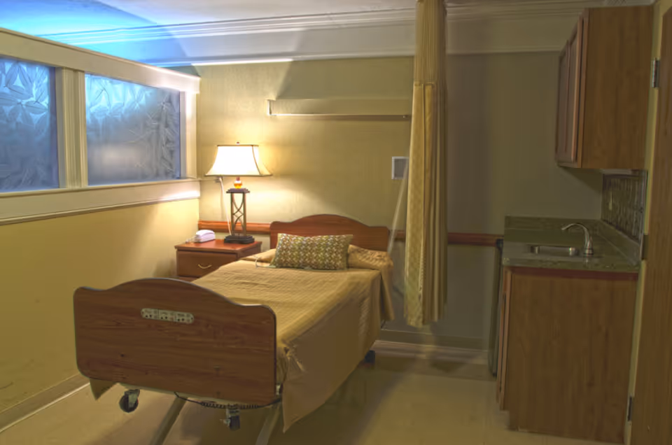 A small, simple room with a single hospital-style bed covered with a beige blanket and a patterned pillow. Next to the bed is a wooden nightstand with a lamp and a telephone. The room has a window with frosted glass, a curtain partially drawn, and a small kitchenette area with a sink and wooden cabinets.