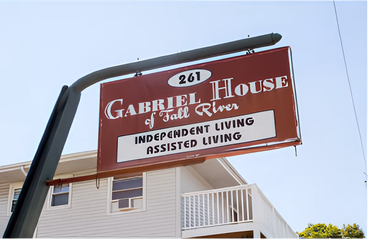 Red hanging sign reading "Gabriel House of Fall River - Independent Living Assisted Living" in front of a light-colored building with a balcony.