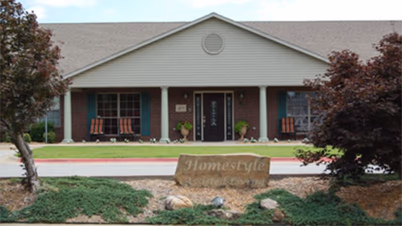 Front entrance of a single-story assisted living building with a covered porch, columns, rocking chairs, and a stone sign reading 'Homestyle Assisted Living'.