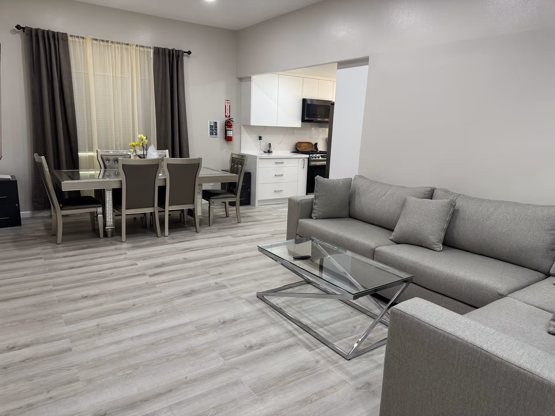 A modern living and dining area with light wood flooring. The living room features a gray sectional sofa with matching cushions and a glass coffee table with a chrome base. The dining area has a glass-top table with six gray upholstered chairs. In the background, there is a small kitchen with white cabinets, a microwave, and a stove. A window with sheer and dark curtains is visible behind the dining table.