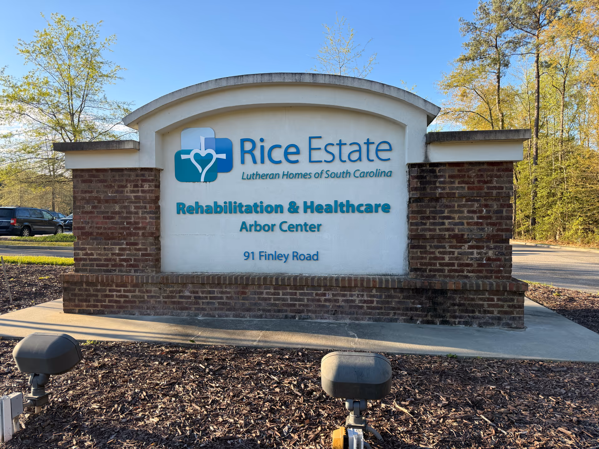 Outdoor sign reading Rice Estate Rehabilitation & Healthcare Arbor Center, with brick base and surrounding trees.