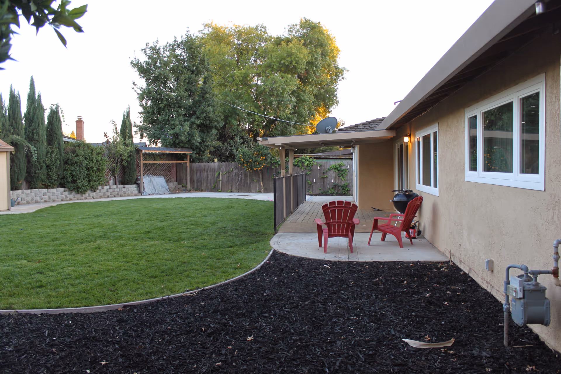 A backyard area with a green lawn, a concrete patio with two red chairs, a small charcoal grill, and a covered porch attached to a beige building. There are trees and bushes along the wooden fence in the background.