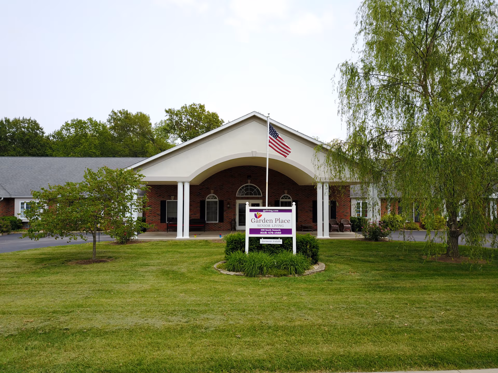 Front exterior view of Garden Place Millstadt senior living facility with a brick building, white columns, an American flag on a flagpole, and a sign in front on a well-maintained lawn with trees.