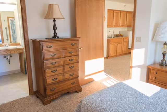 A bright bedroom in an assisted living facility featuring a wooden chest of drawers with a lamp on top, a bed with a patterned bedspread, and an open door leading to a kitchen area with wooden cabinets. A bathroom sink and mirror are visible through another open door.