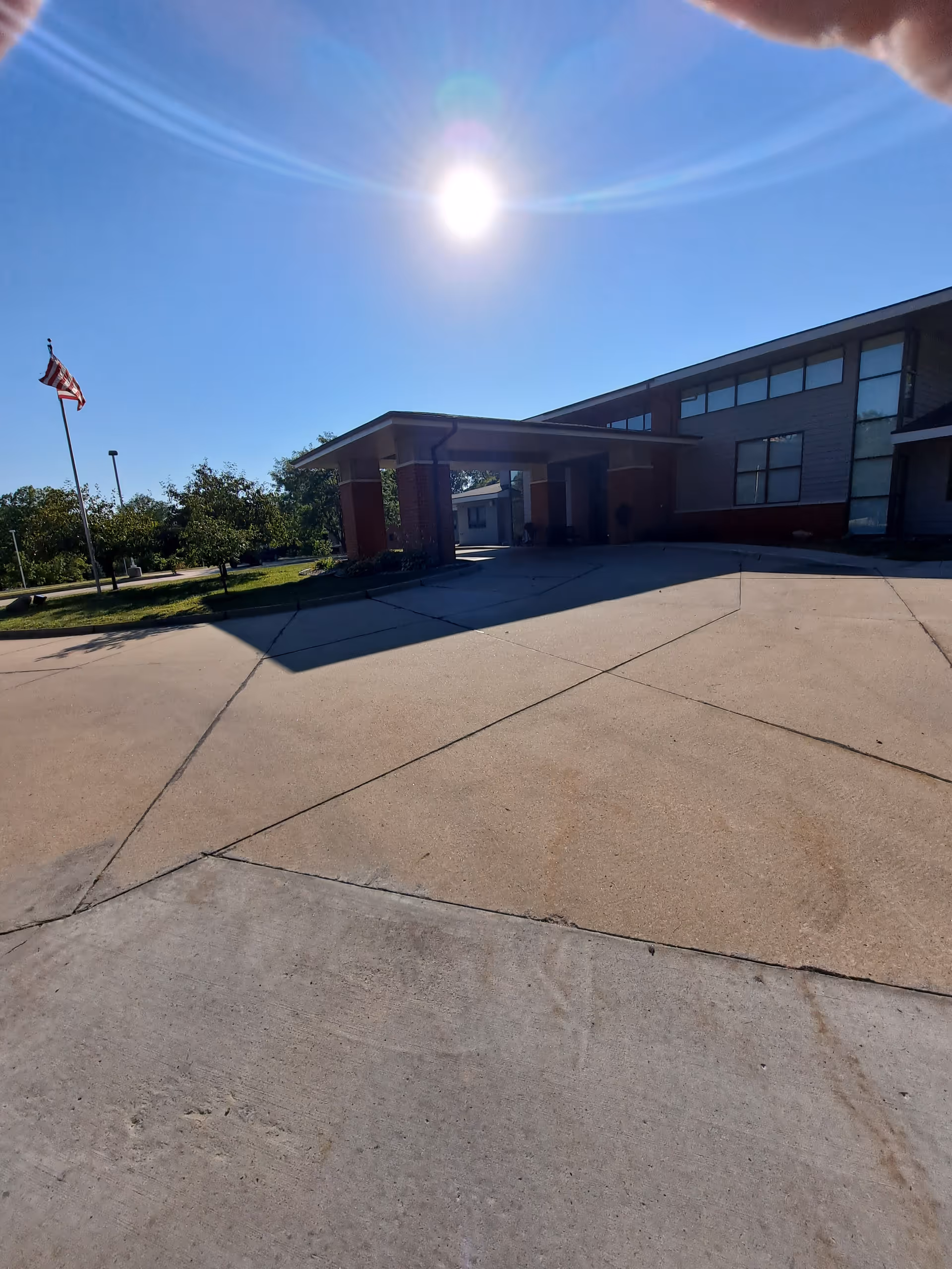 Sunny exterior view of a senior living facility entrance with flagpole and driveway.